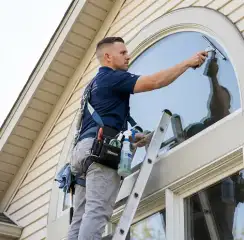 Professional window cleaner on a ladder using a squeegee to clean an exterior arched window, Fargo, ND.